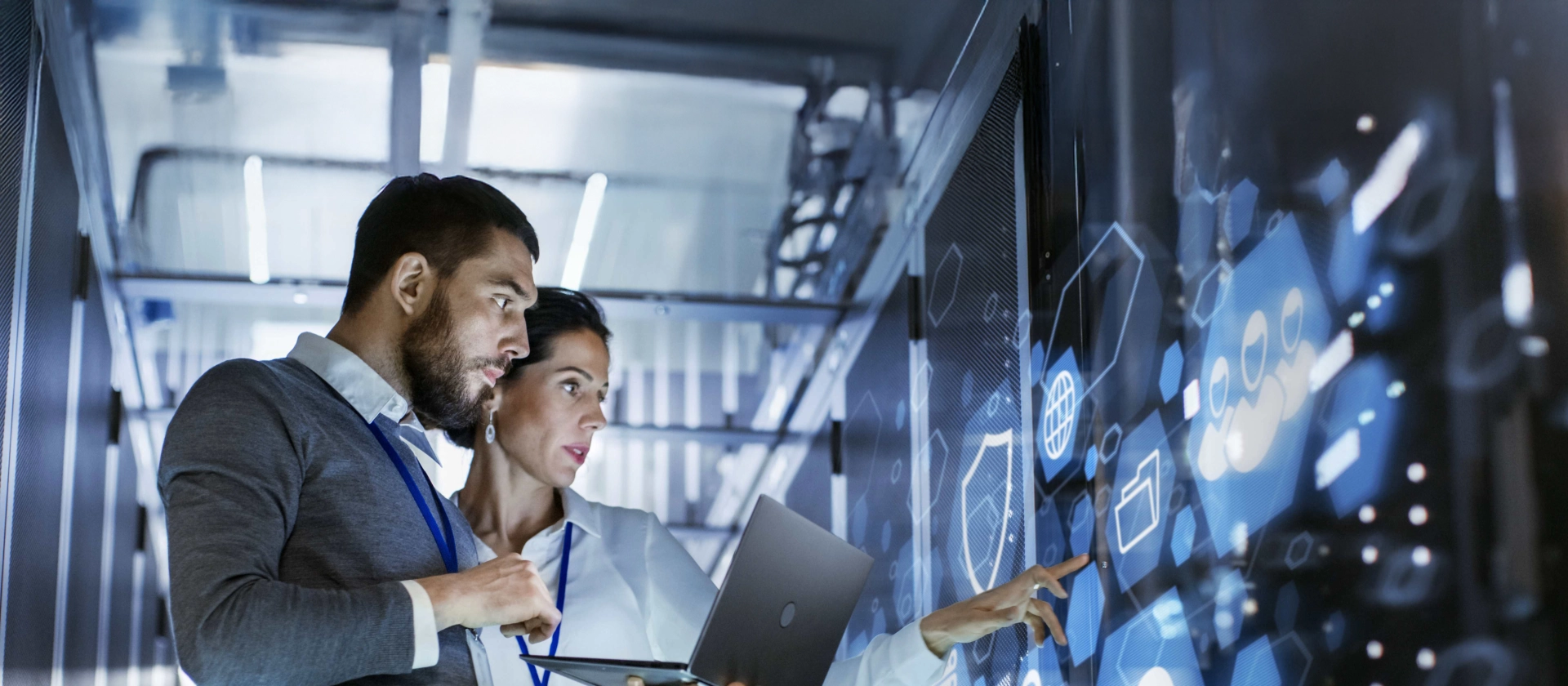 Zwei IT-Fachkräfte in einem Serverraum analysieren Daten auf einem holographischen Display mit Cloud- und Sicherheitssymbolen.