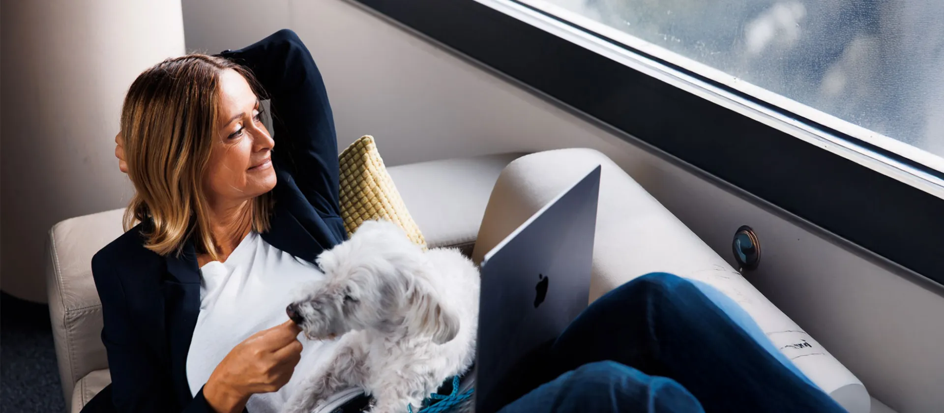 Woman working remotely with laptop and small white dog on lap, enjoying relaxed atmosphere by the window.