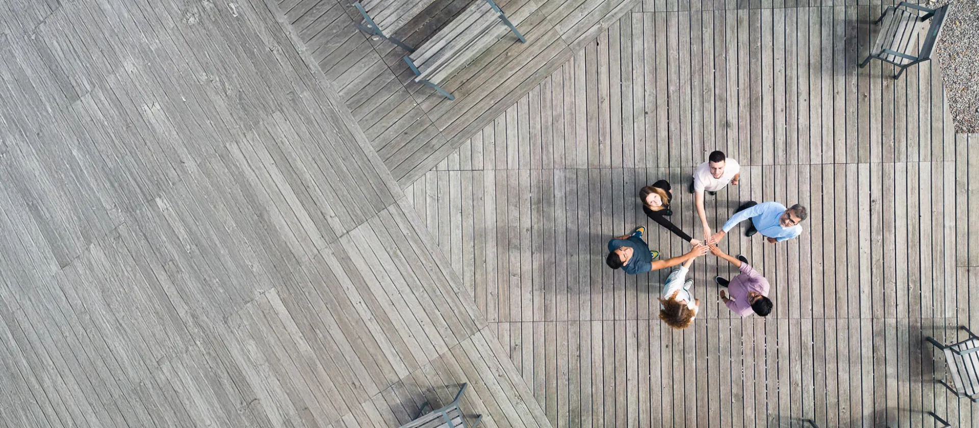 Aerial view of a diverse business team standing in a circle on a wooden deck, joining hands in the center to symbolize collaboration, innovation, and shared goals.