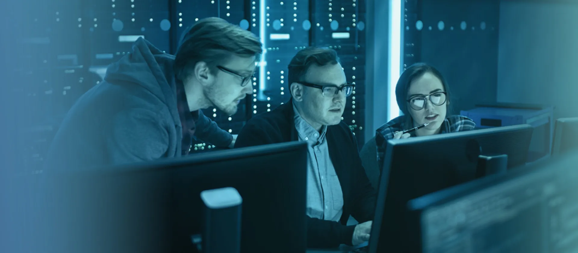 Zwei Kollegen und eine Kollegin aus dem Bereich Cyber Security sitzen konzentriert an einem Arbeitsplatz, von blauem Gradient überlagert