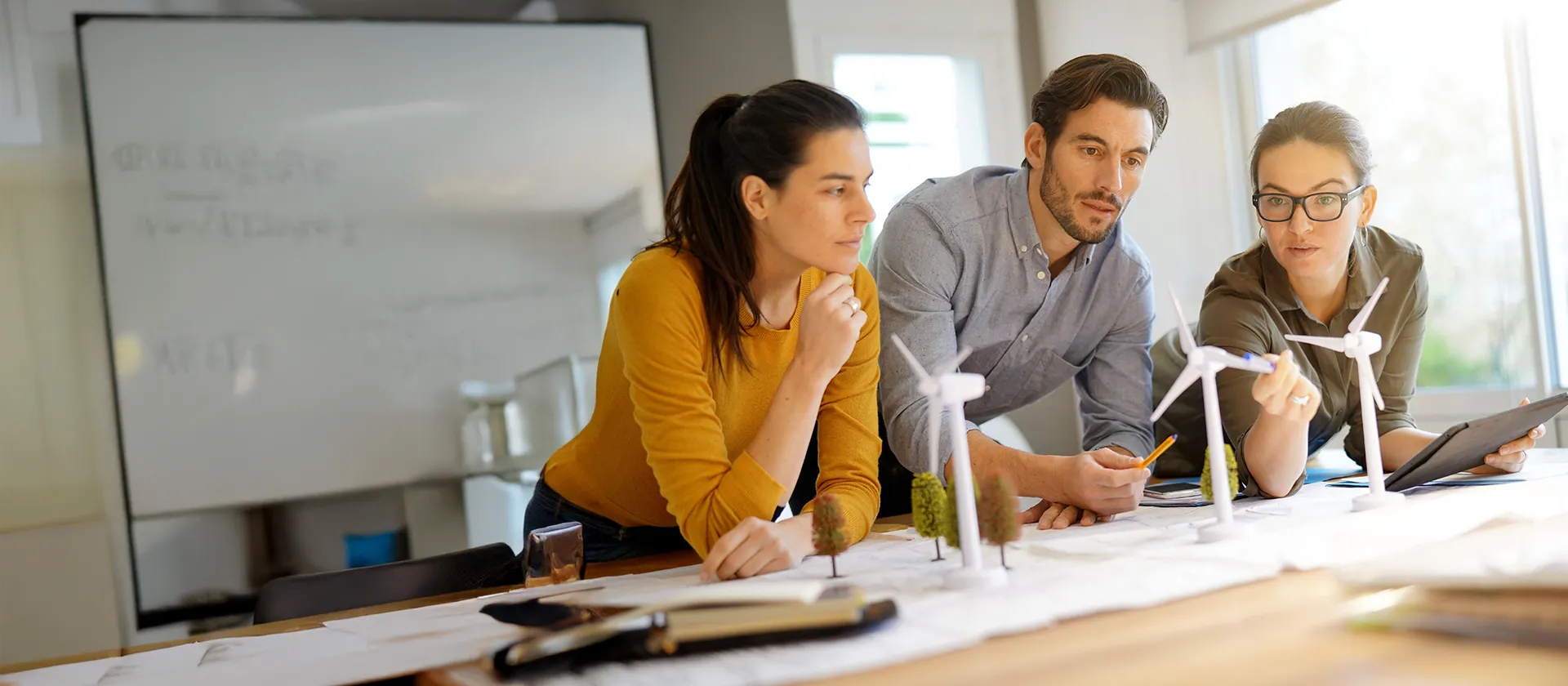 Three professionals collaborating on a wind energy project, analyzing miniature wind turbines and sustainability concepts in a bright, modern office environment.