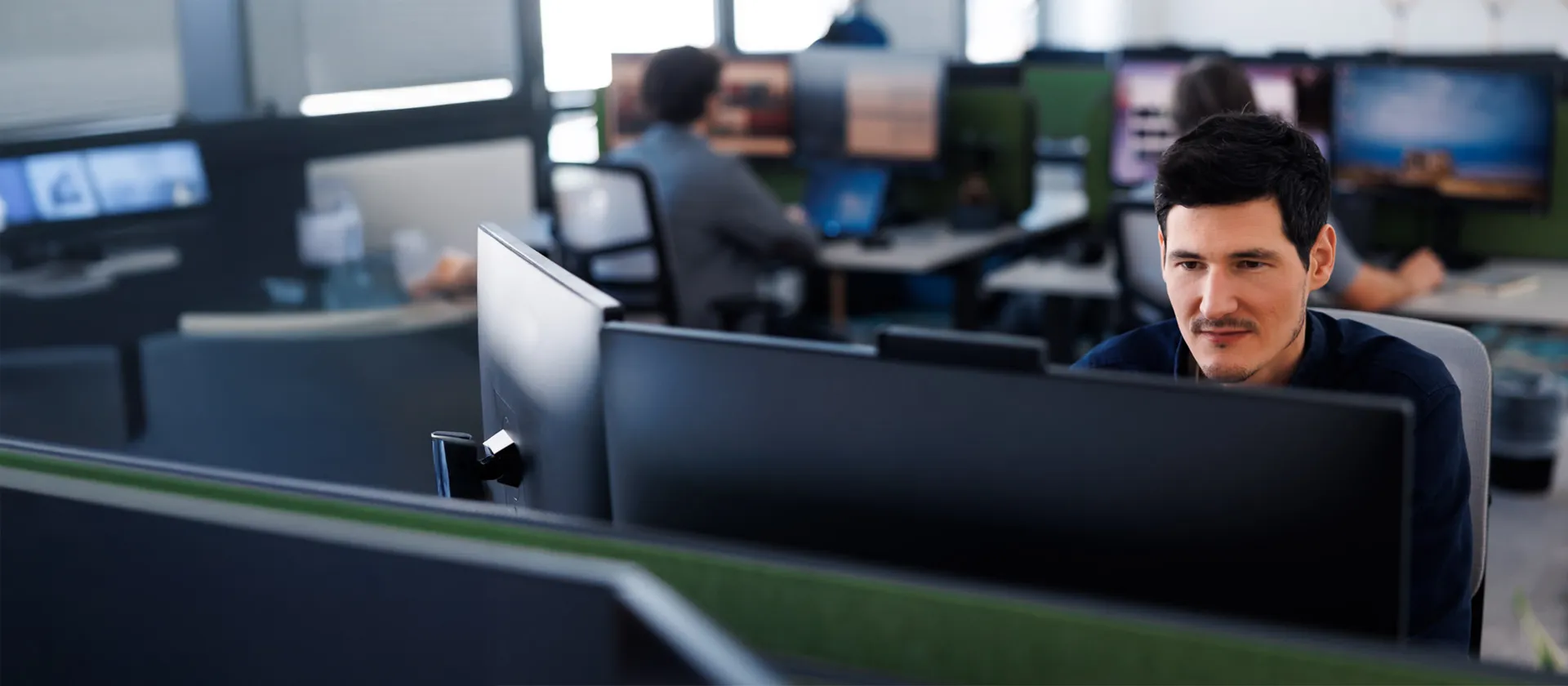Male IT professional working intently on a dual-monitor setup in an open office environment, representing software development, SAP services, or digital operations.