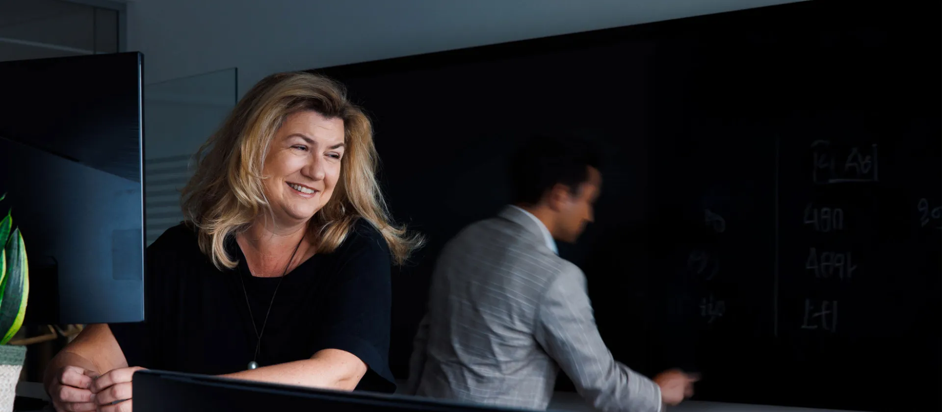 Smiling woman at a desk with a male colleague writing on a blackboard in the background at msg Plaut office.