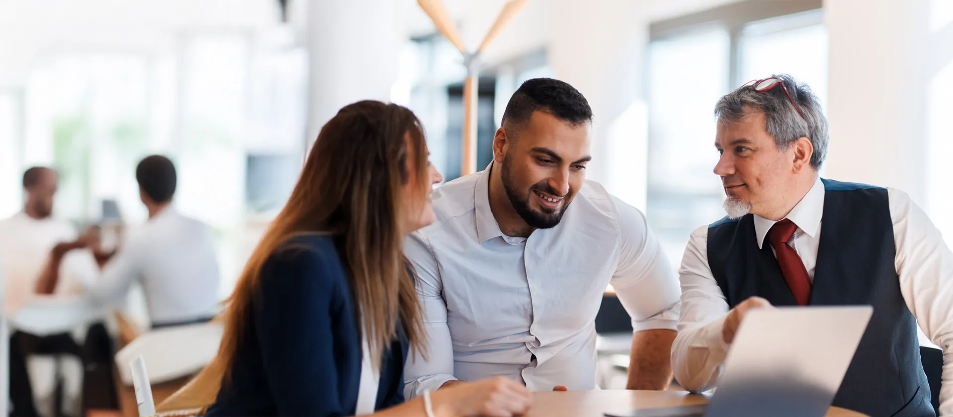 Three professionals in a collaborative discussion around a laptop in a bright office setting, symbolizing teamwork, consulting, and digital transformation planning.
