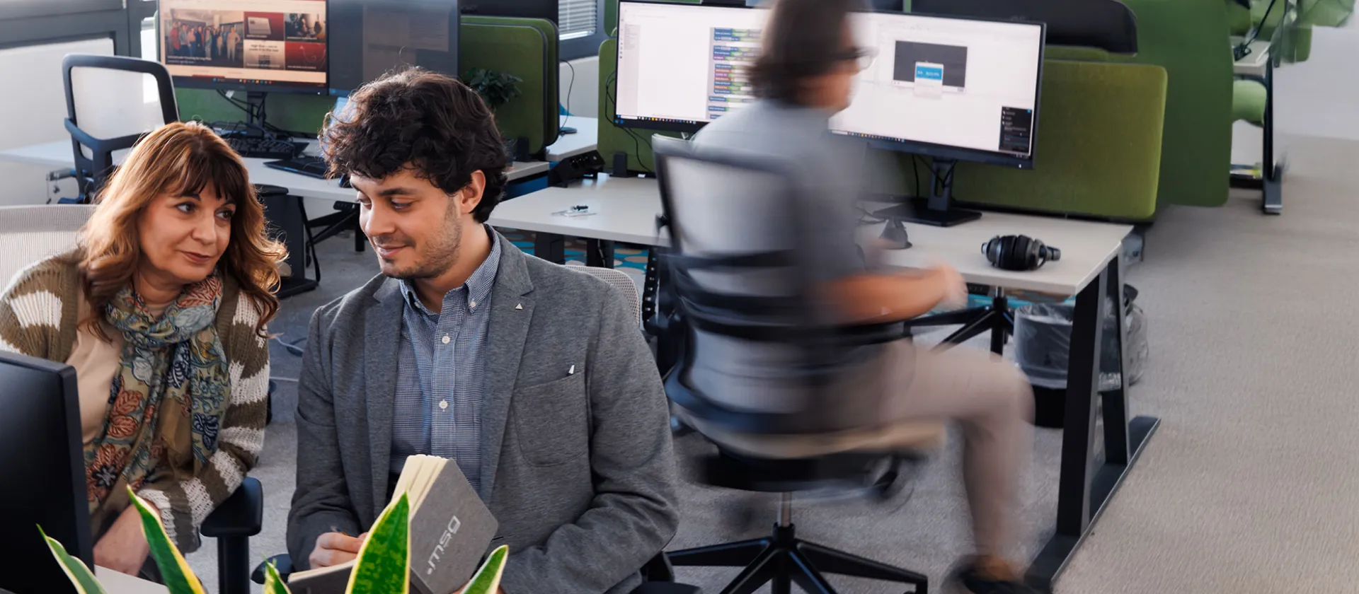 wo colleagues in a modern office discussing a project while reviewing notes on a laptop, with an open-plan workspace in the background.