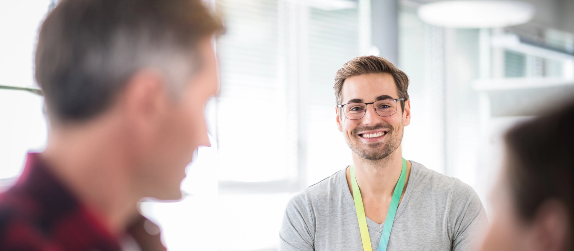 Nachwuchsentwickler lächelt inmitten einer Bürosituation über die Bildschirme hinweg in die Kamera