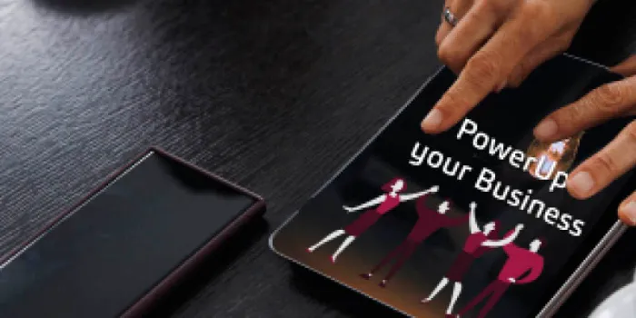 Tablet screen with “Power Up Your Business” text and illustrated team of people celebrating, hand interacting with the screen on a dark wooden table.