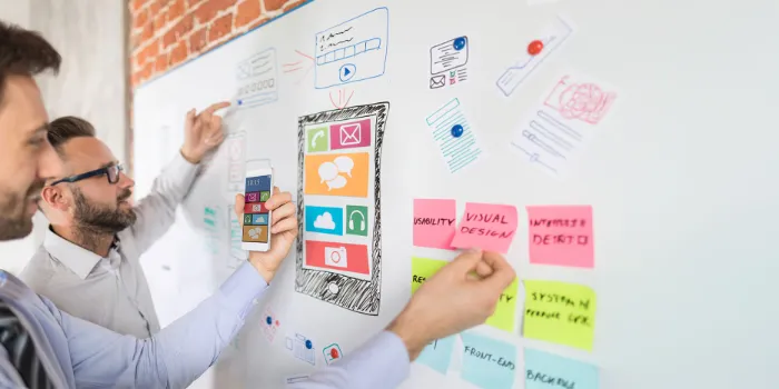 Two UX designers collaborate on a mobile app interface by sketching and organizing user interface elements on a whiteboard, highlighting usability and visual design.