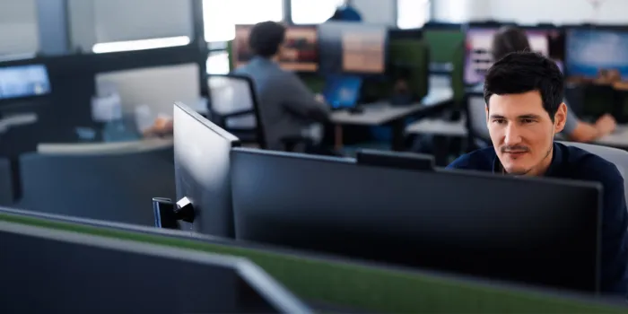 Male IT professional working intently on a dual-monitor setup in an open office environment, representing software development, SAP services, or digital operations.