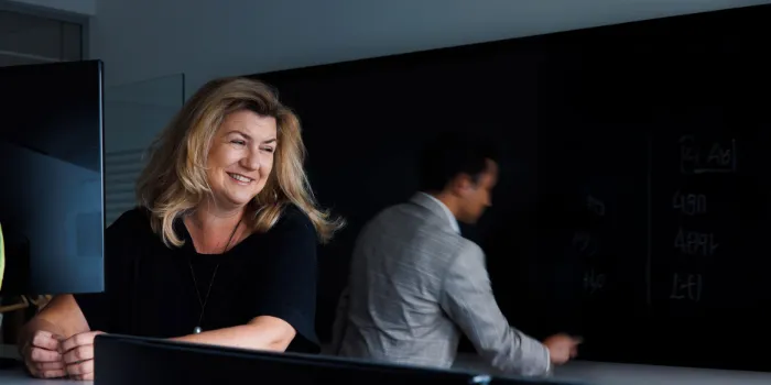 Smiling woman at a desk with a male colleague writing on a blackboard in the background at msg Plaut office.