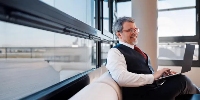 Confident senior executive working on a laptop in a modern office lounge with large windows, showcasing flexible work culture and leadership approachability.