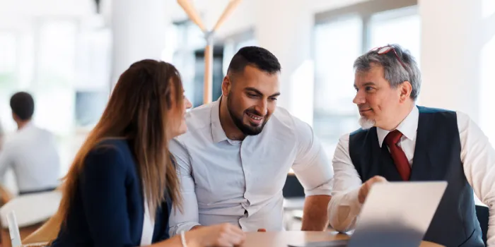 Three professionals in a collaborative discussion around a laptop in a bright office setting, symbolizing teamwork, consulting, and digital transformation planning.