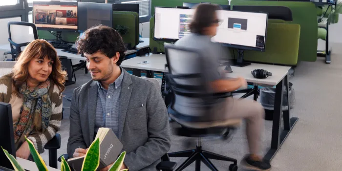 wo colleagues in a modern office discussing a project while reviewing notes on a laptop, with an open-plan workspace in the background.