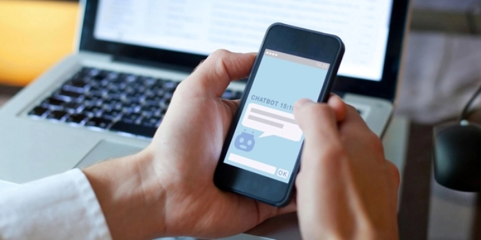 Mensch hält mit zwei Händen Smartphone mit Chatbot in der Hand, im Hintergrund ein LLaptop