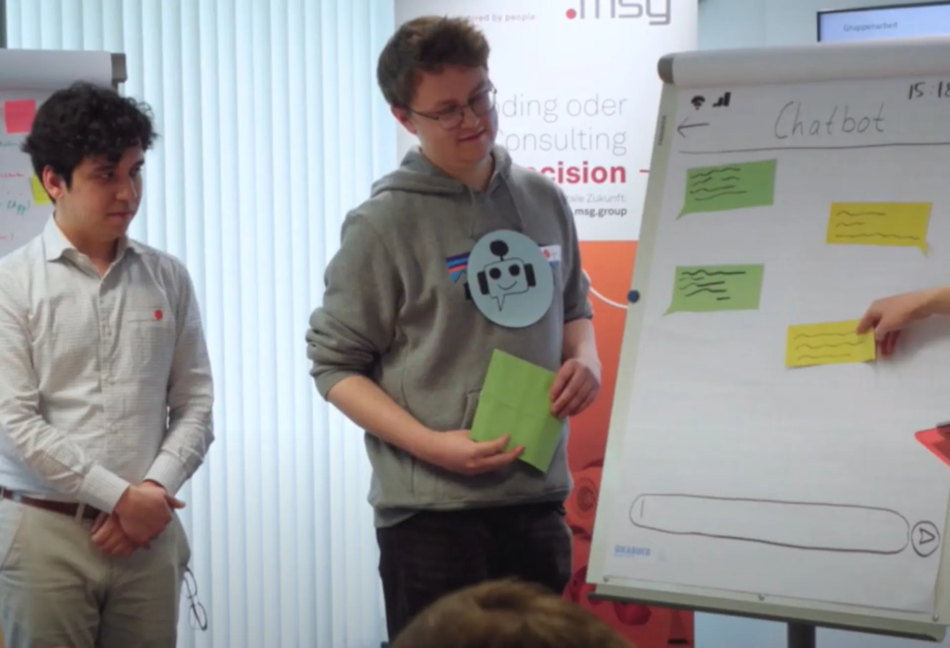 Zwei Studenten stellen an einem Flipchart mit mehreren bunten Zetteln der Gruppe ein Arbeitsergebnis vor